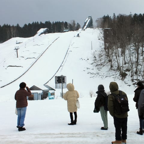 migakiba nayoroのイメージ写真。フィールドワーク訪問中の写真です。雪舞う中で、フィールドツアーの参加者が、ピヤシリスキー場のすぐそばにあるスキージャンプ台を眺めています。