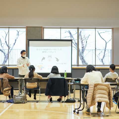 migakiba gojomeのイメージ写真。五城目小学校でのコンポスト授業の様子を撮影した画像。前のスクリーンにコンポストの説明が投影され、子供たちがメモを取りながら聞いている。
