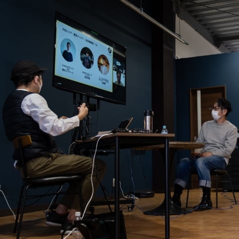 migakiba oaraiのイメージ写真。現地報告会の会場ARISEにて、ゲストコメンテーターの笹目亮太郎さんと現地事務局代表の鈴木さんがモニターに映されたZoomの画面を前に、コメントしている様子が写っています。