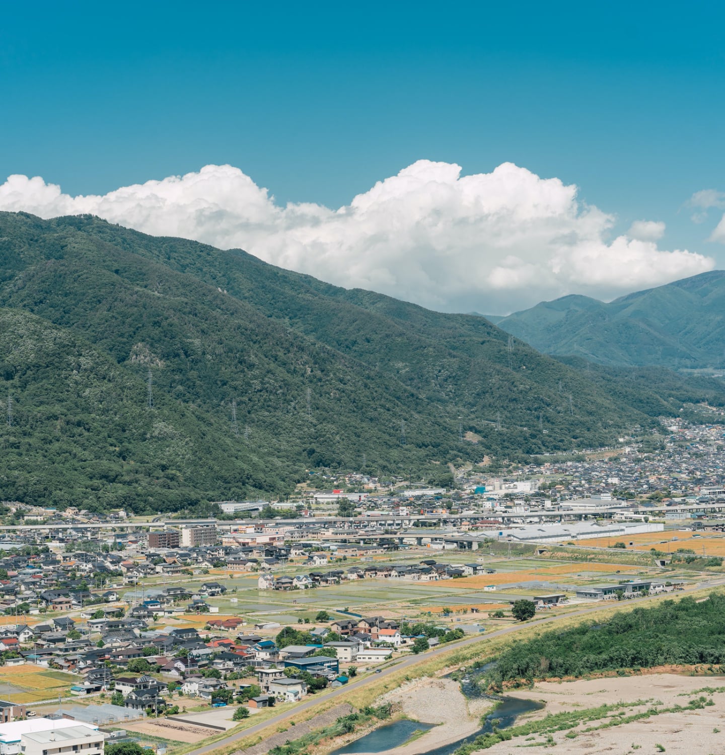 写真：migakiba uedaの上田市街地。背景には、上田市の北側にそびえ、上田のシンボルともいわれる「太郎山」の山並みが広がっています。手前の近景には北陸新幹線が走っており、線路を挟むように住宅、商業施設、田畑があり、街並みを形成しています。
