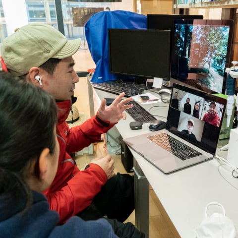 写真：migakiba yamaguchiで、実施した現地成果報告会の様子。YCAMバイオリサーチラボで、現地事務局代表の菅沼さんと、現地チームの高原さんがパソコンの画面に向かい話しています。