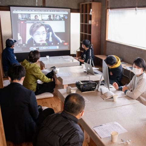 写真：migakiba gotoで実施した現地成果報告会の様子。オンラインで参加したチームのメンバーがプレゼンテーションする様子を大画面のスクリーンに映し出し、大きな机をはさんで、7名の現地事務局メンバーや参加者の方々が聞いています。