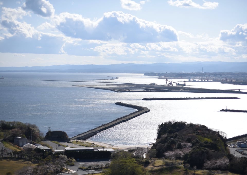 写真：migakiba iwakiのいわき市三崎公園港の見える丘より望む小名浜港。開かれた海の水面に反射する太陽の光と青空の青が混じり合う。右手に小名浜の工業地帯が見えます。