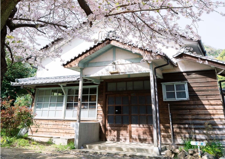 migakiba kureのイメージ写真。現地事務局・一般社団法人まめなの拠点である旧梶原医院の入り口写真です。上部には花が咲いた桜の木、木造の旧梶原医院の様子が写っています。