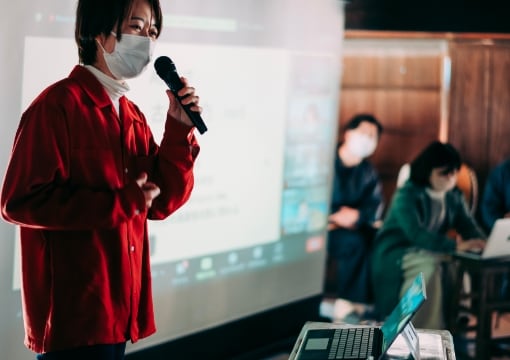 migakiba kureのイメージ写真。現地報告会で名寄参加チームがマイクを持ち、スライドの前で話している様子が写っています。