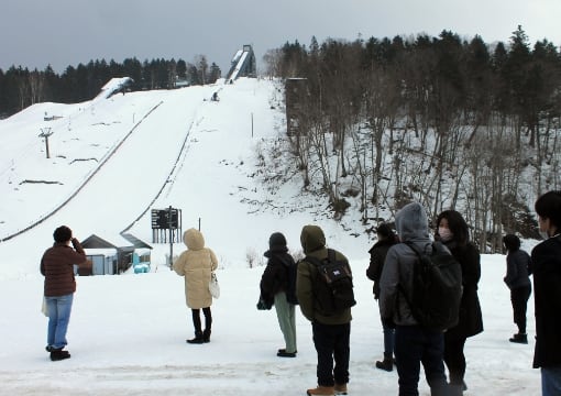写真：migakiba nayoroのイメージ写真。フィールドワーク訪問中の写真です。雪舞う中で、フィールドツアーの参加者が、ピヤシリスキー場のすぐそばにあるスキージャンプ台を眺めています。