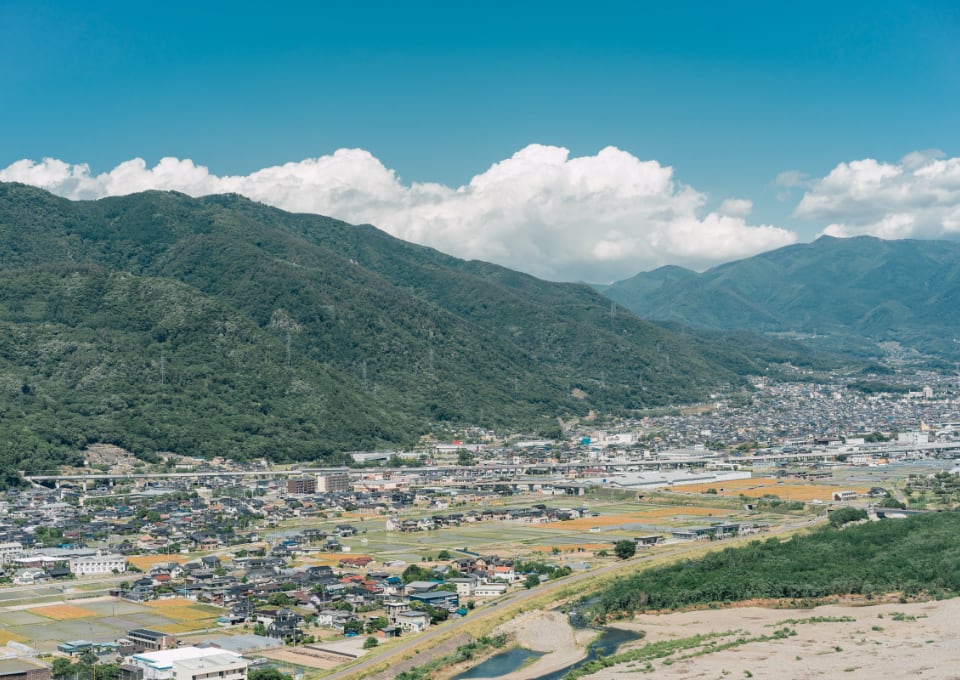 写真：migakiba uedaの上田市街地。背景には、上田市の北側にそびえ、上田のシンボルともいわれる「太郎山」の山並みが広がっています。手前の近景には北陸新幹線が走っており、線路を挟むように住宅、商業施設、田畑があり、街並みを形成しています。