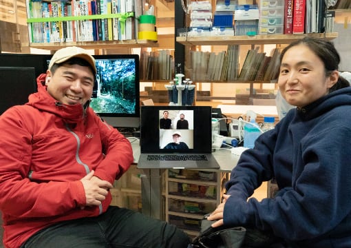 写真：migakiba yamaguchiで、実施した現地成果報告会時の現地事務局・現地チームの記念撮影。YCAMバイオリサーチラボにいる、現地事務局代表の菅沼さんと、現地チームの高原さんに挟まれたパソコンの画面にはオンラインで参加しているその他の現地事務局・現地チームのメンバーがうつっています。
