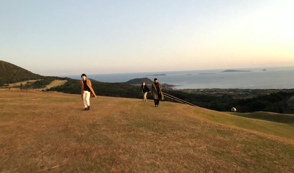 写真：migakiba gotoで実施したフィールドツアーの様子。夕暮れの五島の鬼岳の写真です。手前に茶色の芝生の丘、奥の方に海や山が見渡せます。現地事務局の中村さん、現地アドバイザーの有川さんが頂上に向かって登ってきています。