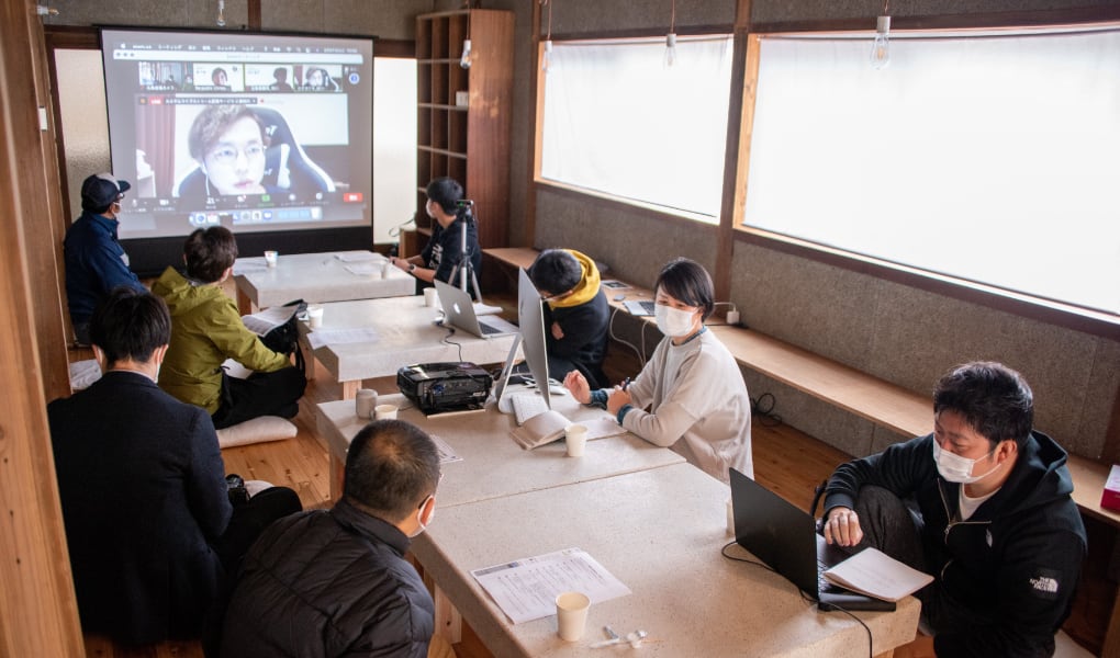 写真：migakiba gotoで実施した現地成果報告会の様子。オンラインで参加したチームのメンバーがプレゼンテーションする様子を大画面のスクリーンに映し出し、大きな机をはさんで、7名の現地事務局メンバーや参加者の方々が聞いています。