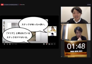 PCキャプチャ画像：migakiba goto 代表チームのプレゼンテーションの様子。スライドが投影されており、2名のプレゼンターとタイムキープの時計が映っています。