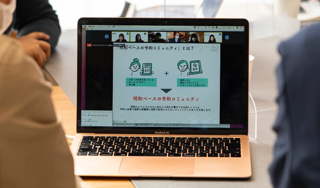 写真：migakiba kashibaの現地報告会の様子。PC画面に参加チームのプレゼンテーションが映っており、それを会場から見ている様子が映っています。