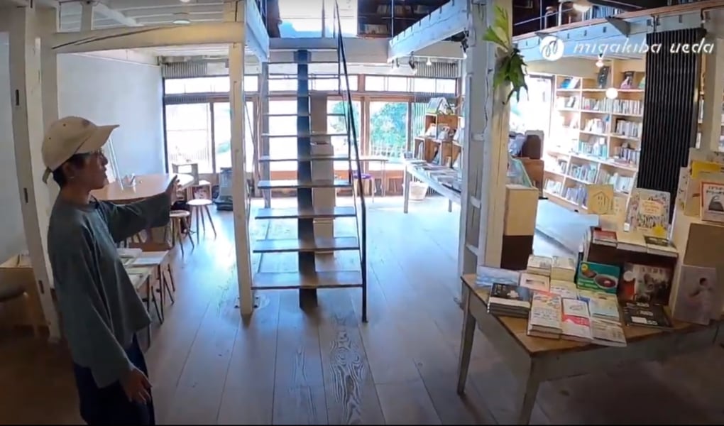 写真：migakiba uedaで実施したフィールドツアーの様子です。現地事務局バリューブックスの実店舗、本屋未満の店内です。木のぬくもりが感じられる明るい吹き抜けの店内に、新館を中心とした本が棚やテーブルに置かれています。その様子を、事務局長の池上さんが案内しています。
