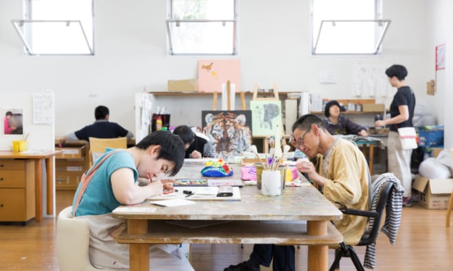 写真：migakiba kashibaのフィールドワーク先候補「アートセンターHANA」の建物内部。障がいを持つ複数のメンバーがそれぞれの創作活動をしています。テーブルに向かっている2人は絵の具や色鉛筆など様々な画材をつかい真剣なまなざしで絵を書いています。