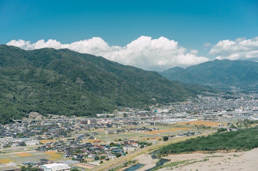 写真：migakiba uedaのロゴと上田市街地を見渡した風景。背景には、上田市の北側にそびえ、上田のシンボルともいわれる「太郎山」の山並みが広がっています。手前の近景には北陸新幹線が走っており、線路を挟むように住宅、商業施設、田畑があり、街並みを形成しています。