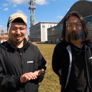 写真：migakiba yamaguchiの担当者2名。カジュアルな服装で屋外に立ち、笑顔でカメラを向いている。